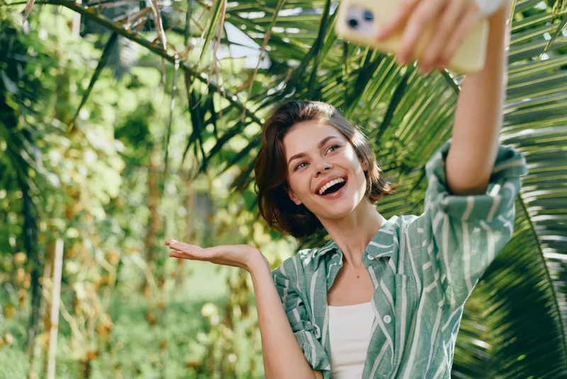 Woman enjoying a sunny outdoor moment, a cheerful portrait as she smiles for a selfie amid lush greenery. Casual fashion, relaxed pose, vibrant mood in nature.