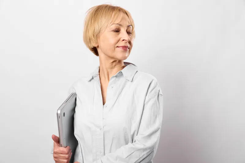 Senior woman blonde portrait in white shirt, holding notebook, thoughtful expression, standing against gray background, neutral studio light, calm mood, looking to side