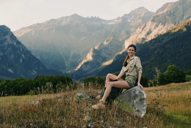 Young woman sits on a rock in a grassy meadow with distant mountains and a clear sky, enjoying a peaceful outdoor moment during a sunny day.