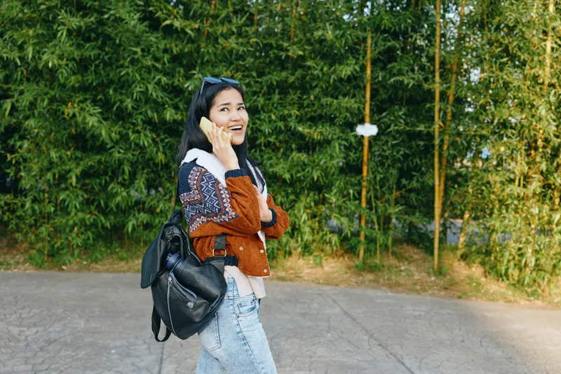 Woman talking on phone outdoors with backpack, casual jacket and jeans along a forest path, smiling under sunny daylight, urban vibe meets nature in a lifestyle photograph for travel and communication
