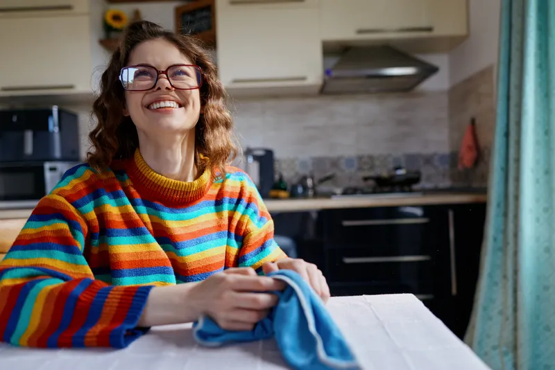 A cheerful woman in a striped sweater sits at a kitchen table, ironing a cloth in a bright home setting. A candid lifestyle moment highlighting domestic chores and comfort.