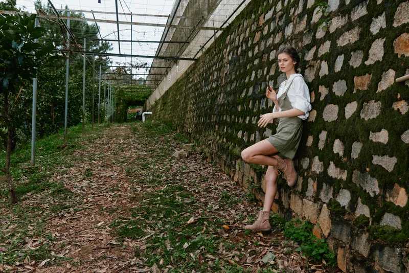 Woman in casual outfit posing against a stone wall in a lush garden, showcasing a serene expression and natural surroundings for lifestyle photography
