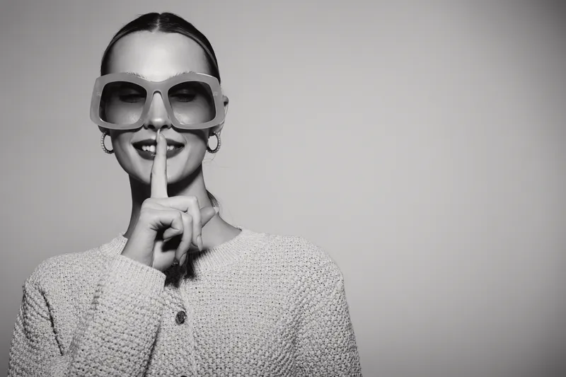 Young white woman with fair skin wearing sunglasses and a knitted sweater makes a shushing gesture with finger on lips. Concept of silence, secrecy, and calm expression.