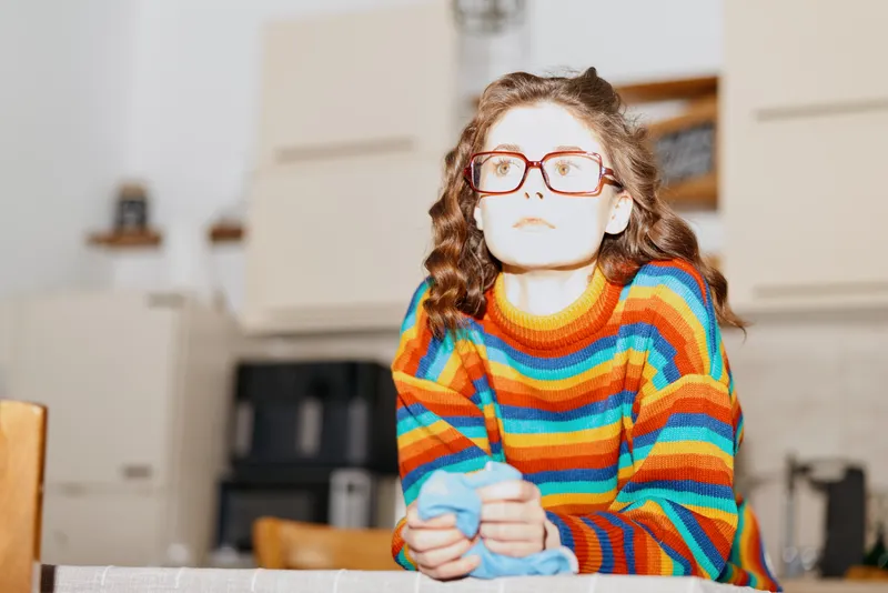 A woman at home sits at a table in a bright kitchen, wearing a striped sweater and glasses, gazing upward with a thoughtful expression. Concept: home life, calm, introspection.