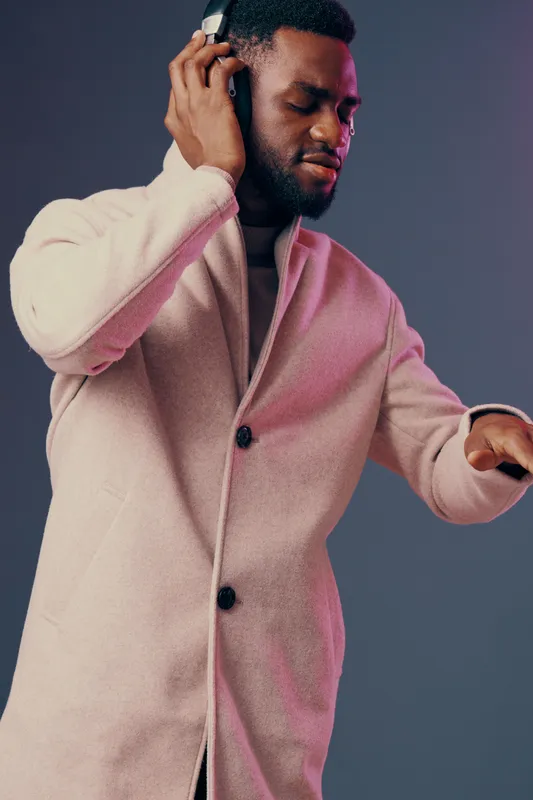 Stylish young man enjoying music in a pink coat with headphones, expressing joy and relaxation against a soft gray background