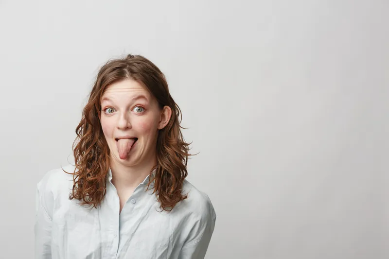 Young woman with curly hair wearing white shirt sticks out tongue playfully, isolated on light background. Concept of fun, humor, and casual lifestyle portrait.