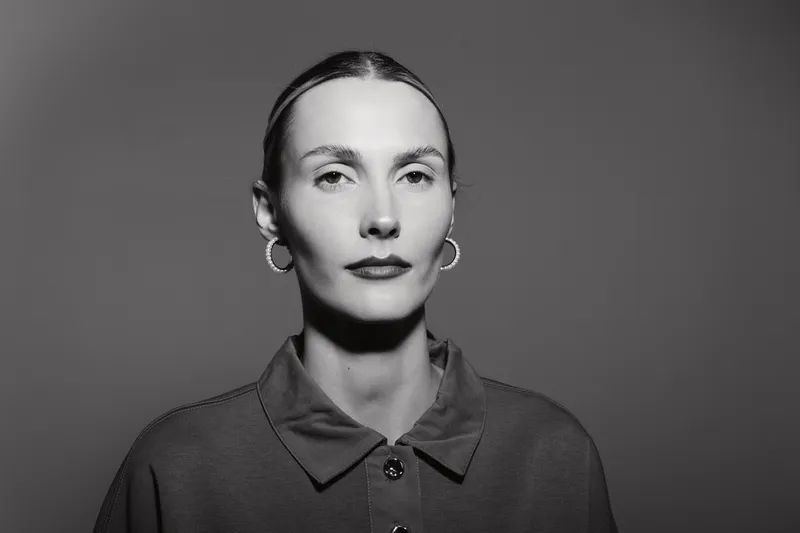Portrait of a serious white woman with pale skin and slicked back hair wearing earrings and a collared shirt on a grey background.