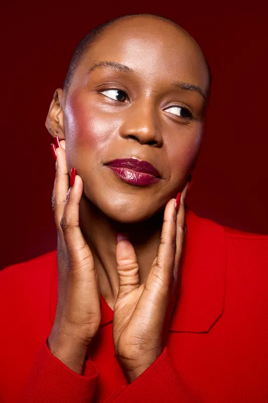Close-up portrait of an elegant black woman with dark skin wearing red jacket and matching red lipstick and nails. She gently touches her face against a red background. Beauty concept.