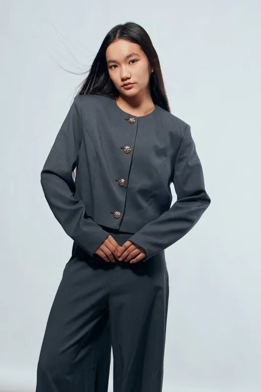 Young woman standing with hands clasped wearing gray formal jacket and matching pants against light background. Portrait, casual business style and confident pose with minimal makeup and long hair.