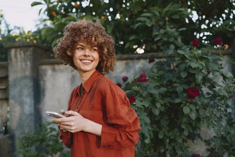 A smiling woman in a red shirt stands in a garden, holding a smartphone. A candid lifestyle moment showing casual confidence and outdoor everyday life