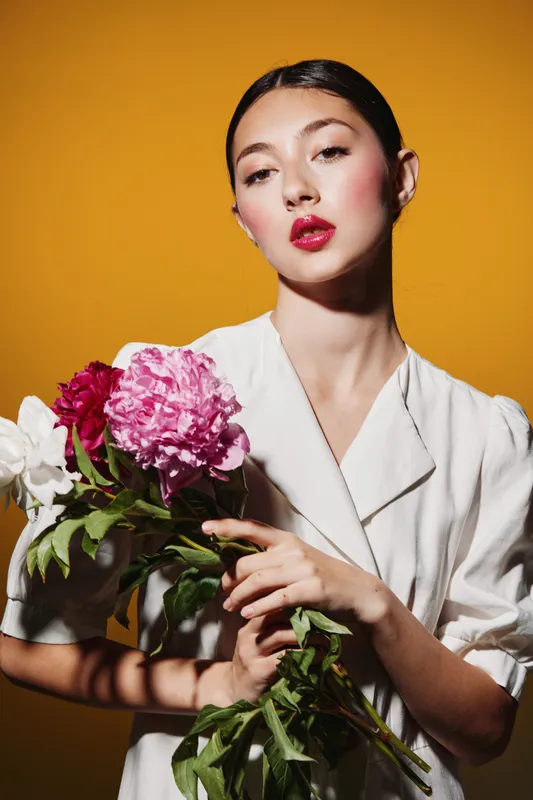 Elegant young woman with glossy dark hair and vibrant lip makeup holding fresh peony flowers on yellow background. Creative fashion beauty portrait with vivid colors studio shot.