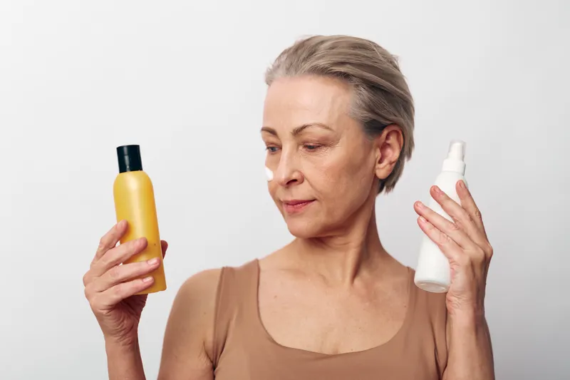 Mature woman with short hair comparing two cosmetic bottles, one yellow and one white, isolated on white background. Concept for skincare choices and personal care routine.