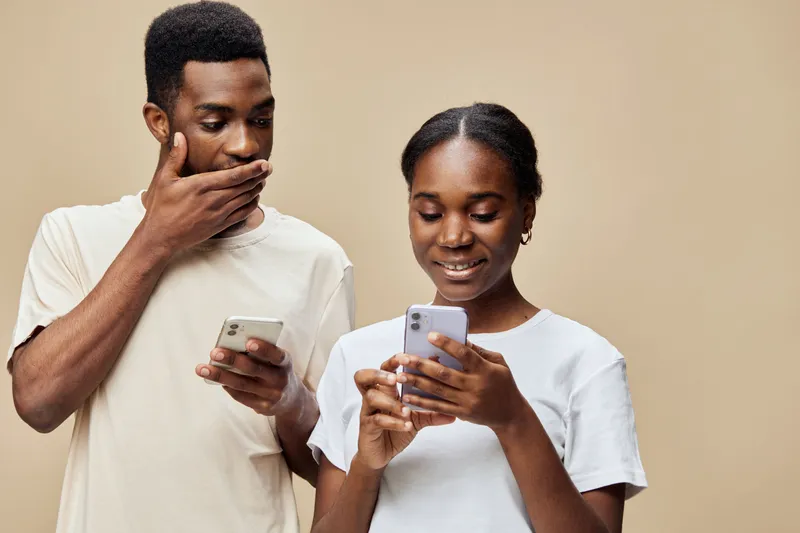 Young interracial couple engaged in social media, displaying emotions of surprise and joy, with neutral beige background creating a warm atmosphere