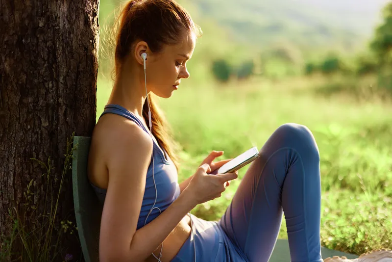 Young woman in athletic clothing enjoying music on a tablet outdoors in nature, conveying relaxation and tranquility during a sunny day