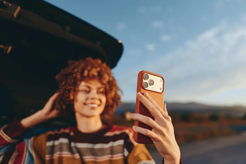Smiling woman with curly hair holding a smartphone for a selfie outdoors, travel lifestyle, open car scene, warm sunset light, clear sky, distant mountains, golden hour glow, coastal road