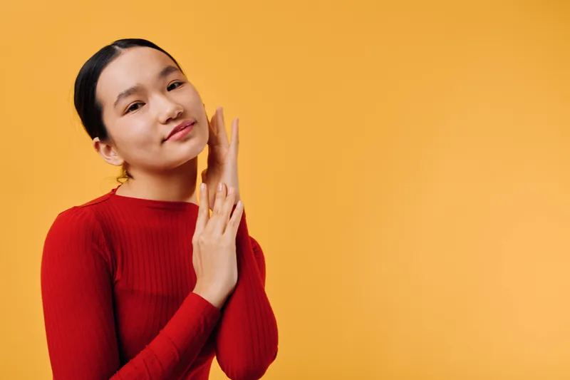 Young woman in red sweater with closed eyes and gentle smile touching her face against orange background. Beauty, calm emotion and relaxed pose with natural light for wellness.