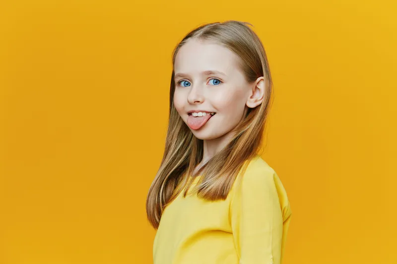 Cheerful girl with blonde hair sticking out her tongue against a vibrant yellow background, showcasing playful emotions and bright fashion