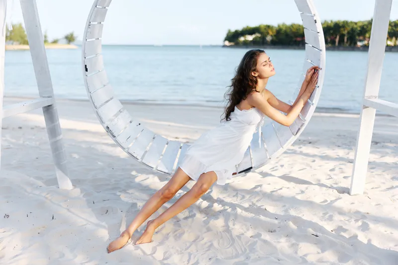 A young woman in a white dress sits on a circular seaside swing, leaning forward with a relaxed pose on bright sand, while soft sunlight and calm blue water create a serene mood.