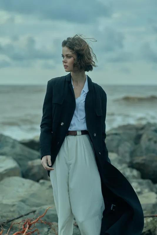 Woman in a long black coat and white shirt stands on a rocky shoreline, wind blowing her hair, coastal fashion scene, ocean breeze, moody outdoor elegance and style