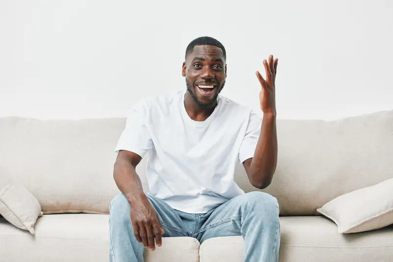 Joyful Black man sitting on a sofa, expressing enthusiasm and engagement, wearing a casual white t shirt and light blue jeans in a minimalist living room