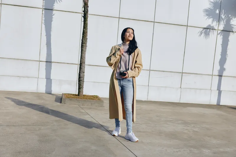 Woman standing outdoors with camera in hand, wearing a long camel coat and jeans beside a white wall and palm shadows, showcasing urban casual style for fashion photography and street wear