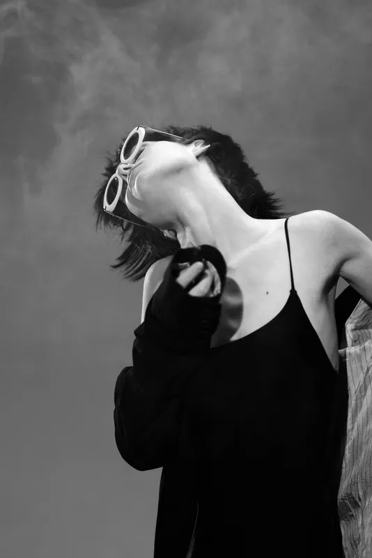 Black and white portrait of a stylish woman wearing sunglasses and a black dress with a dramatic pose against a cloudy sky background.