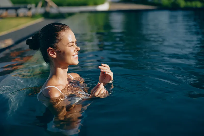 Cheerful woman in a sunlit pool, smiling with eyes closed, relaxed posture, gentle breeze, blue water and lush greenery in the background soft sunlight, clear sky, calm mood, poolside scenic