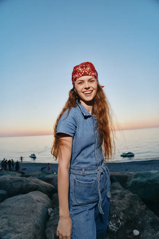 smiling woman standing on rocky beach at sunset wearing red bandana and denim jumpsuit with long hair under clear blue sky