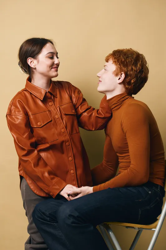 Couple in casual warm clothing sitting and smiling at each other in indoor neutral background, showing affection and connection between young people.