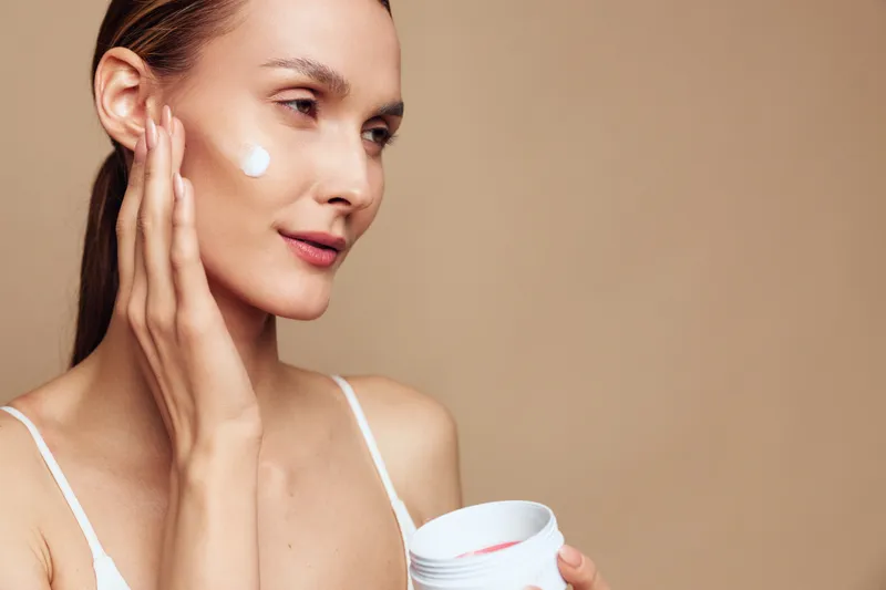 Young woman applies moisturizing cream on face with fingers holding jar of skincare product close to beige background with natural light and smooth skin texture.