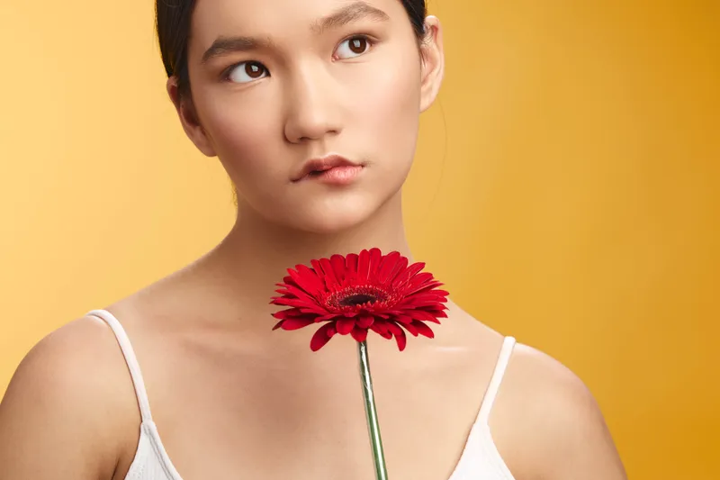 asian woman is seen holding a red flower near her chest against a warm yellow background. She looks contemplative with a soft expression, creating a calm and elegant portrait.