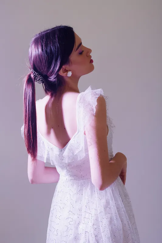 Young woman in a white dress poses with her back turned, showcasing elegant beauty and fashion. Soft lighting highlights her smooth skin and stylish hairstyle. Concept for beauty and fashion.