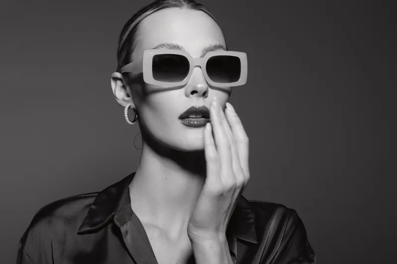 Fashionable white woman with pale skin wearing oversized sunglasses and silk shirt poses with hand near mouth in studio. Glamour and style concept.