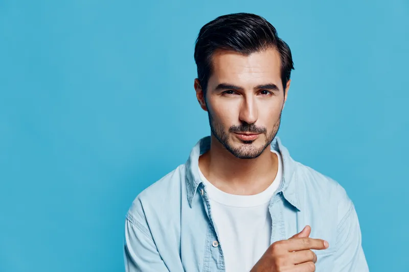 Handsome young man with a stylish haircut wearing a light blue shirt, looking confidently at the camera against a vibrant blue background. Fashion lifestyle concept.