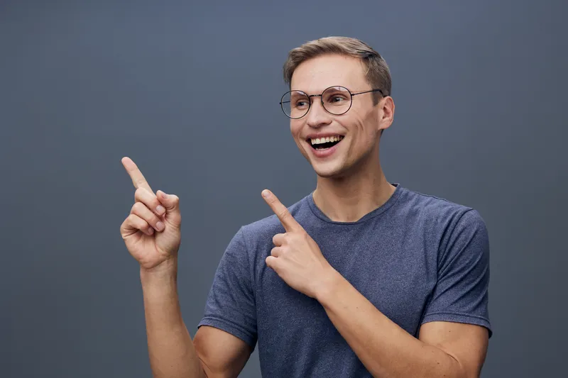 smiling young caucasian man wear glasses blue t-shirt point fingers side look happy studio portrait concept chat AI technology use mobile phone digital communication