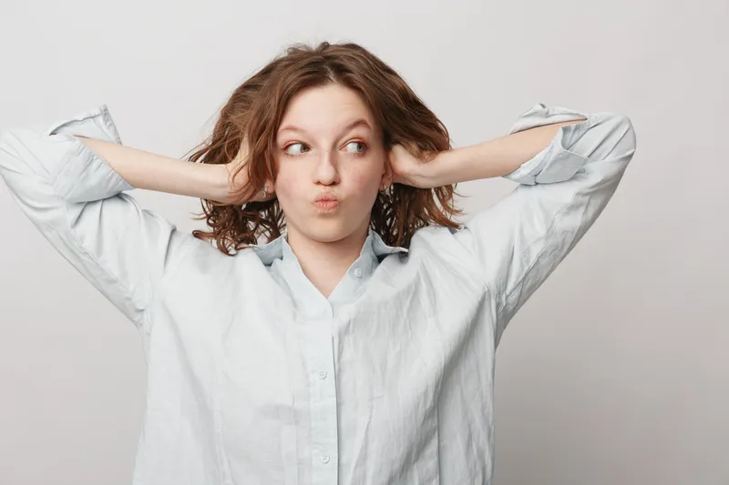 Young woman with curly hair in white shirt covers ears with hands and looks to side. Concept of ignoring noise, stress relief, and playful attitude in casual studio portrait.
