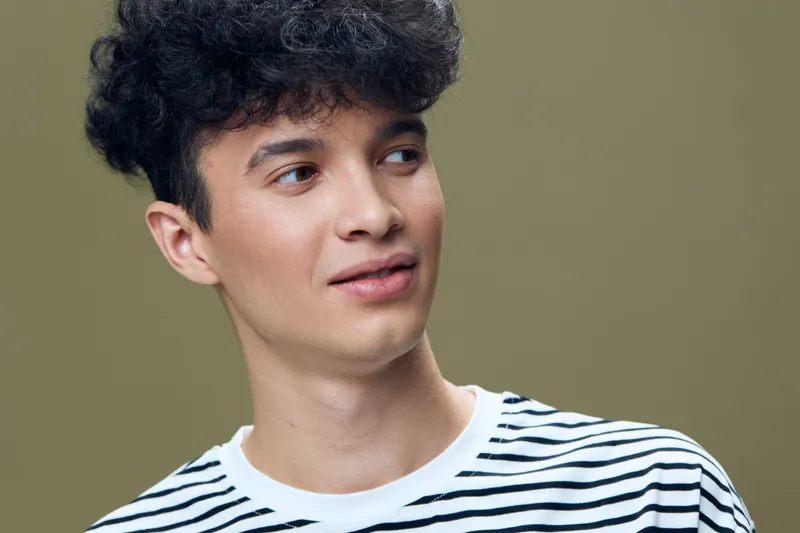 young man wearing a striped shirt with curly hair looking sideways against beige background in natural light