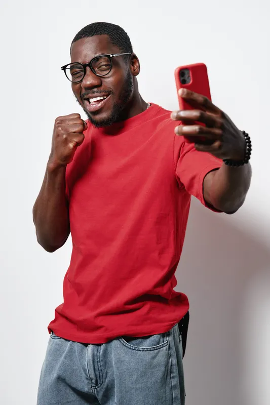 African American man with dark skin wearing glasses and red shirt taking selfie with smartphone, smiling and celebrating success or victory indoors on white background.
