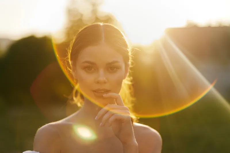 Beautiful young woman in soft sunlight, enjoying nature with a serene expression and gentle pose, wearing a light dress, reflecting summer vibes