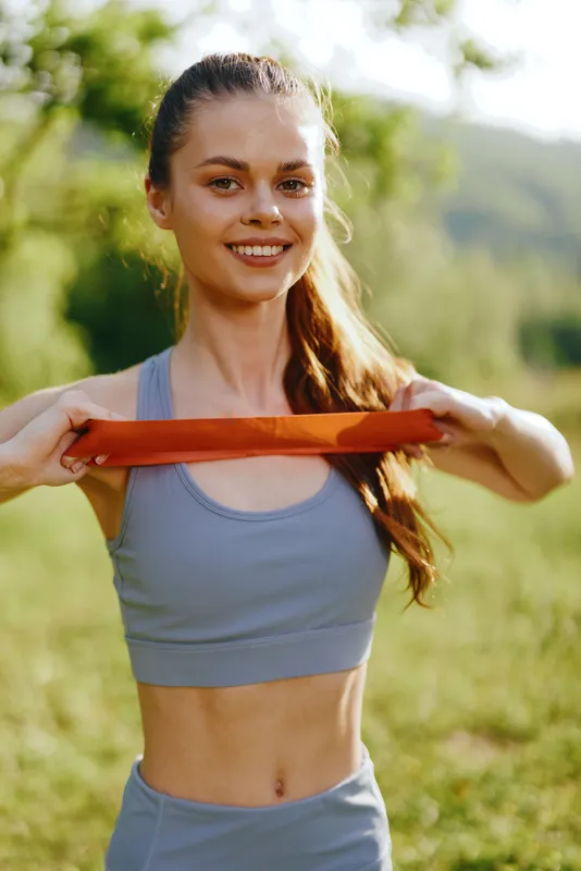 Smiling fitness model using resistance band outdoors in a lush green environment, showcasing healthy lifestyle, strength training, and summer vibes