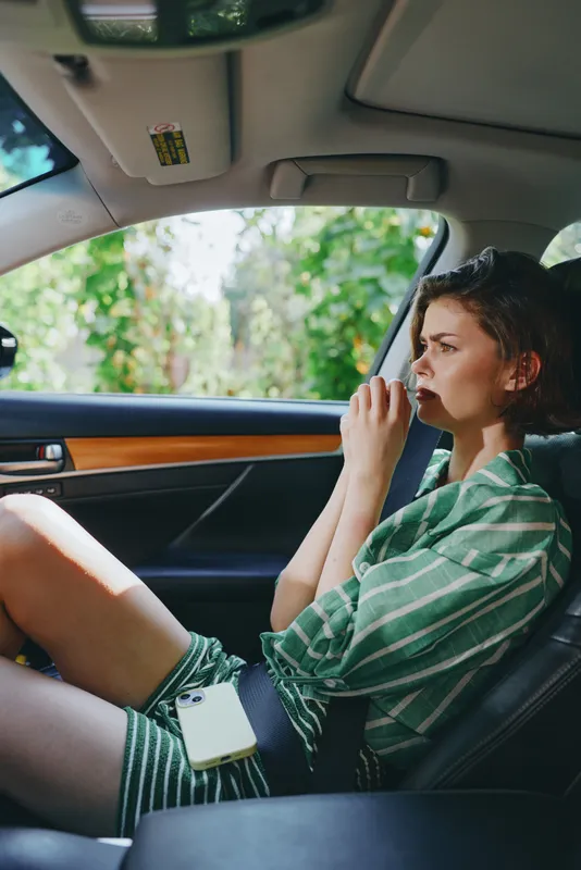 Young woman seated in a car interior, thoughtful gaze, casual green striped shirt, hands near face, sunlit exterior blurred through window.