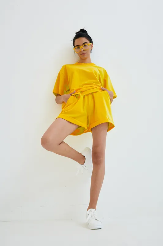 Woman wearing vibrant yellow casual outfit and matching sunglasses stands confidently against white background, showcasing modern summer fashion and relaxed style.