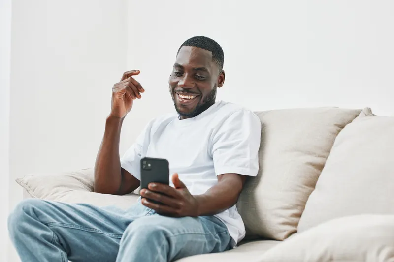 Cheerful African man in casual attire smiling while using a smartphone on a light colored sofa, showcasing happiness and modern lifestyle