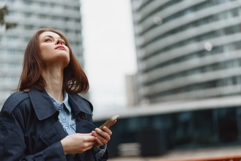 Young brunette woman in a dark jacket looks upward thoughtfully while holding a smartphone in an urban environment with modern buildings in the background.