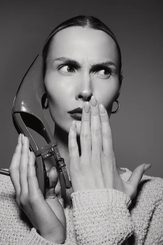 Young Caucasian woman with fair skin holds a high heel shoe near her face, looking puzzled and thoughtful. Concept of fashion, emotions, and footwear.