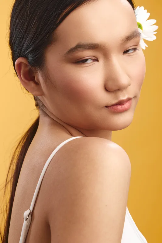 Asian woman posed against a warm backdrop, her delicate features framed by a white strap, a flower tucked behind her ear, conveying grace, poise, and calm in soft light.