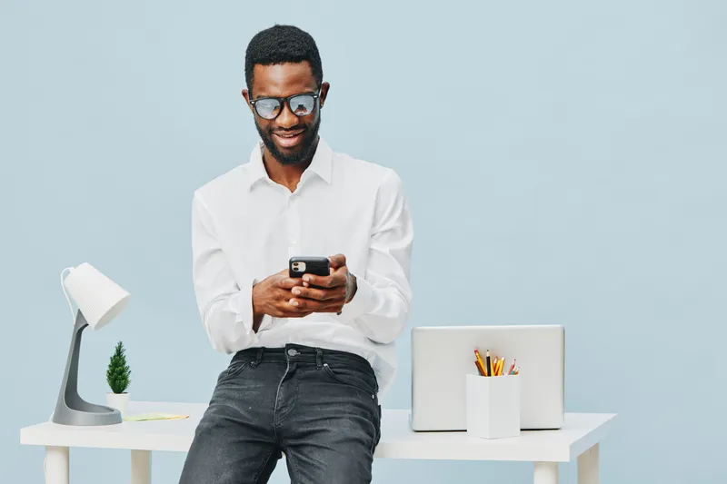 Modern smiling man with glasses in white shirt leaning against desk, using smartphone, with laptop, plant, and stationery on light blue background in a professional workspace. People lifestyle concept