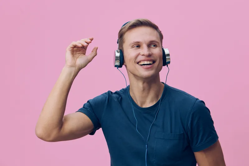 Smiling young man with headphones enjoying music on pink background, casual clothing, leisure activity, happy expression, vibrant colors, studio shot, lifestyle concept.