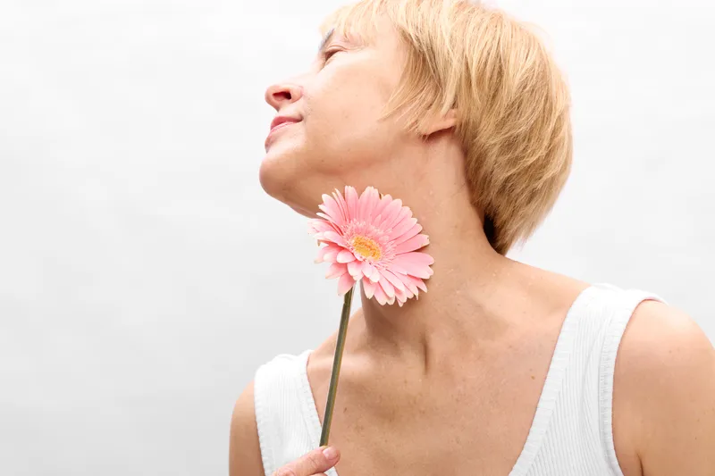 Woman with short hair in white background, holding pink gerbera flower near face, side profile portrait, calm expression, soft natural beauty, minimal style
