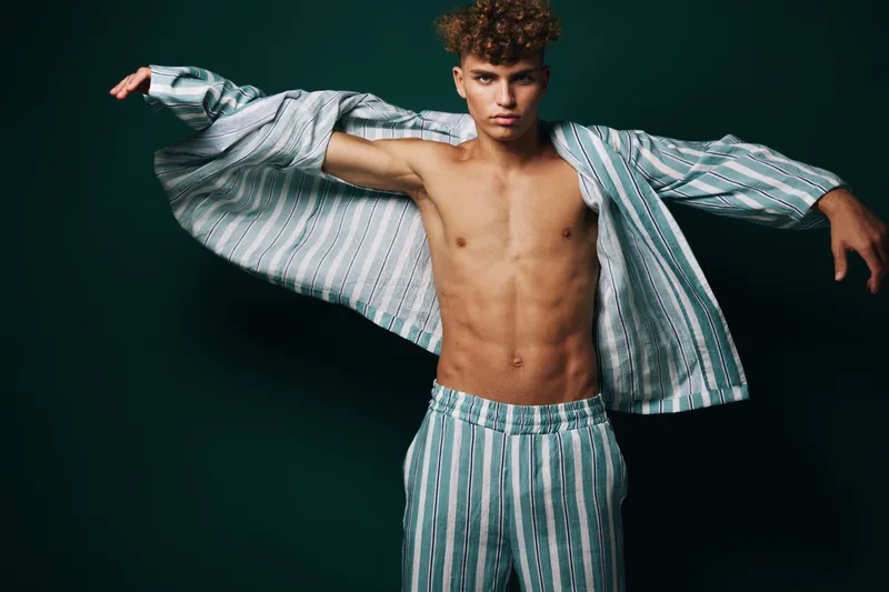 Young man with curly hair wearing striped open shirt and matching pants posing with arms outstretched against dark background in studio. Fashion, style and confidence.